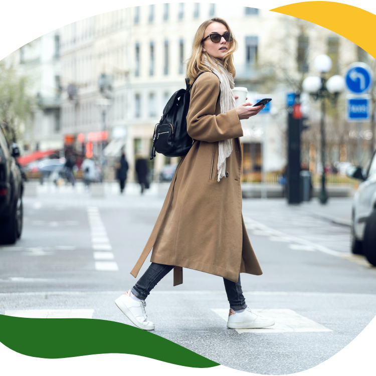 Moderna sieviete ar lielām saulesbrillēm un bēšu garo mēteli šķērso ielu, turot labajā rokā mobilo telefonu