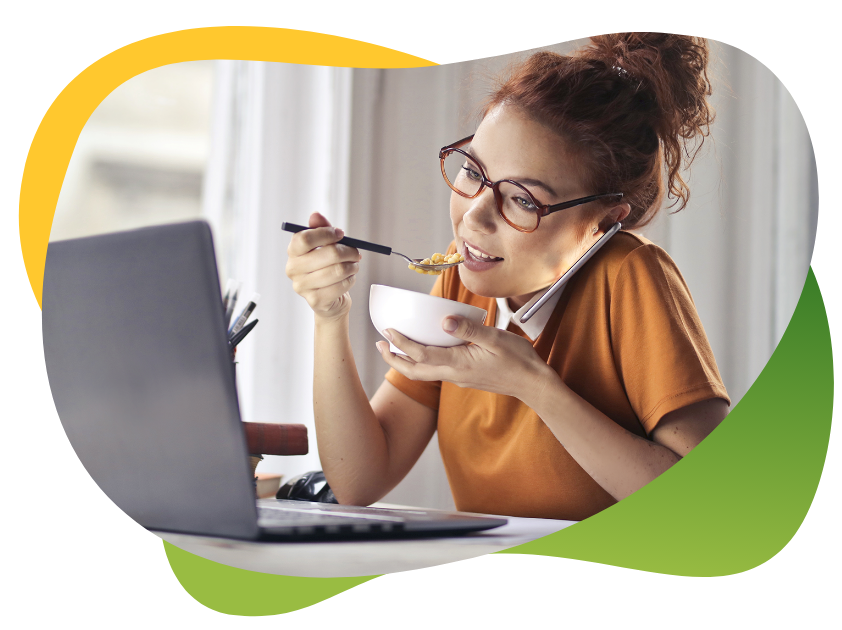 Jauna sieviete ar brillēm un augstu copi sēž pie portatīvā datora, runā pa telefonu un vienlaikus ēd
