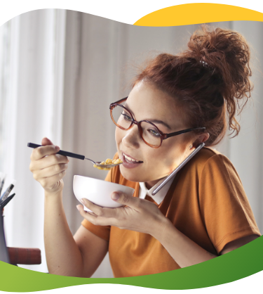 Jauna sieviete ar brillēm un augstu copi sēž pie portatīvā datora, runā pa telefonu un vienlaikus ēd